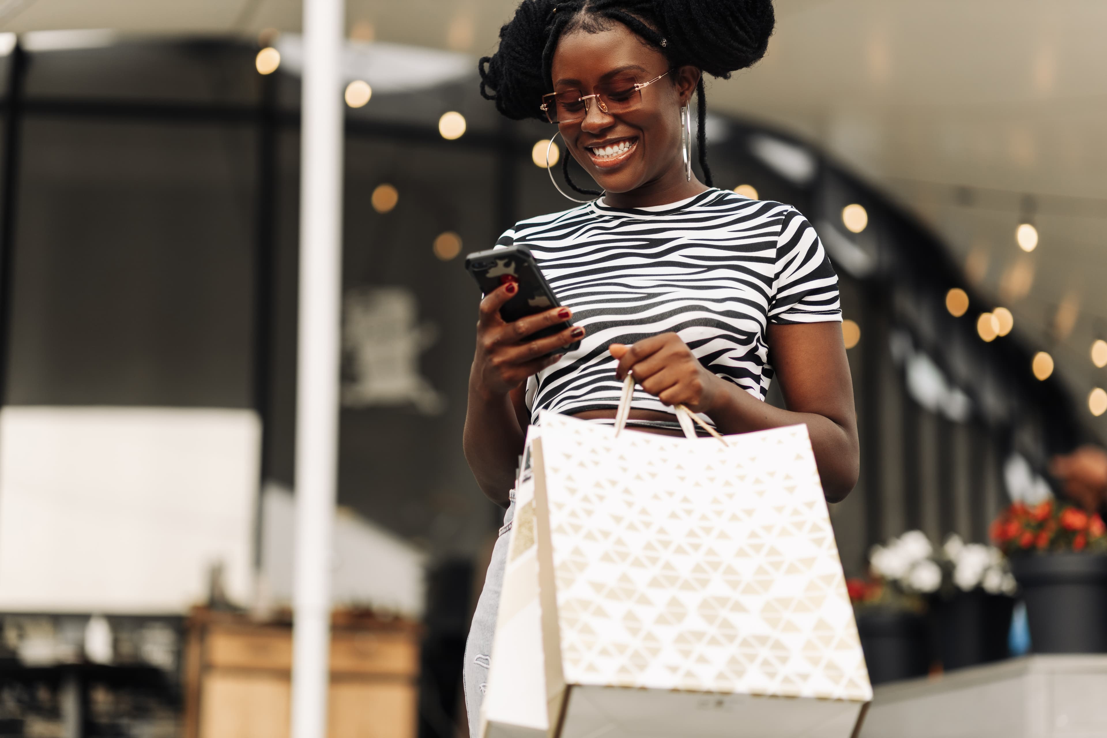 gen z girl holding bags, using smartphone and smiling while shopping